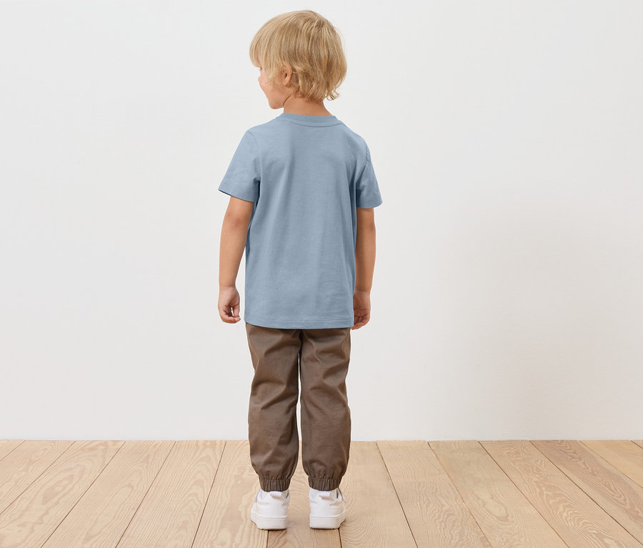 Chłopiec o blond włosach stoi tyłem, ubrany w niebieską koszulkę, brązowe spodnie i białe buty.