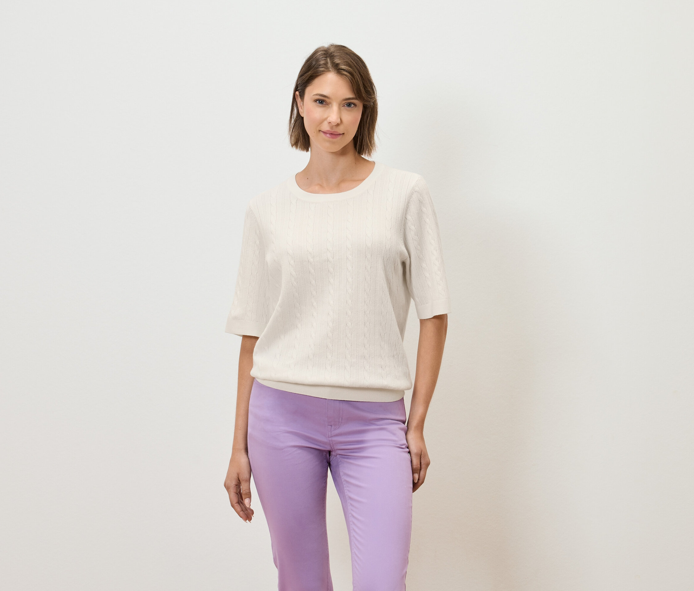 Kobieta pozuje na jasnym tle w kremowym swetrze z krótkim rękawem i fioletowych spodniach.
