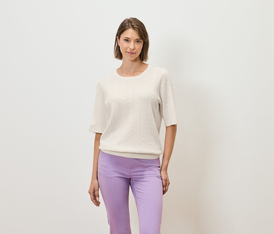 Kobieta pozuje na jasnym tle w kremowym swetrze z krótkim rękawem i fioletowych spodniach.