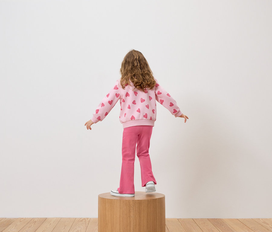 Dziewczynka stoi tyłem do kamery na drewnianym postumencie, ubrana w różową bluzę w serduszka i różowe spodnie.