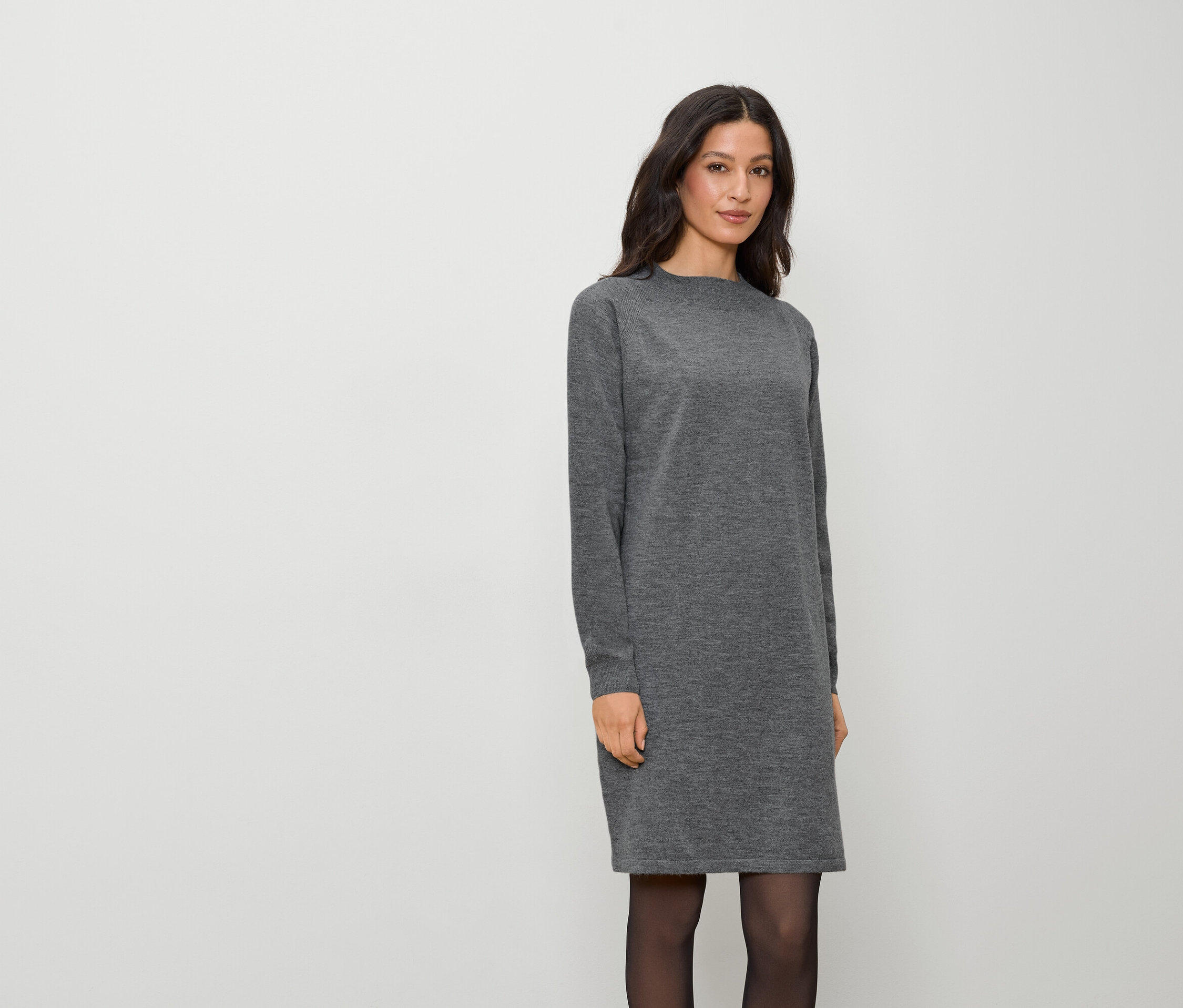 Kobieta w szarej sukience z długim rękawem i czarnych rajstopach stoi na białym tle.