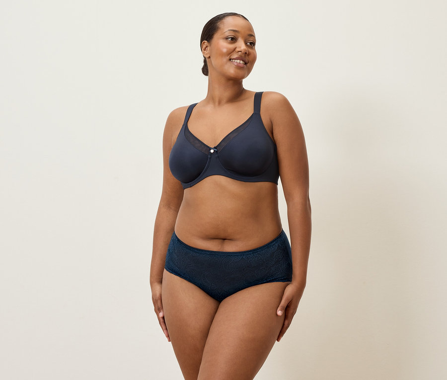 Kobieta w średnim wieku z ciemnymi włosami i granatową bielizną stoi na kremowym tle.