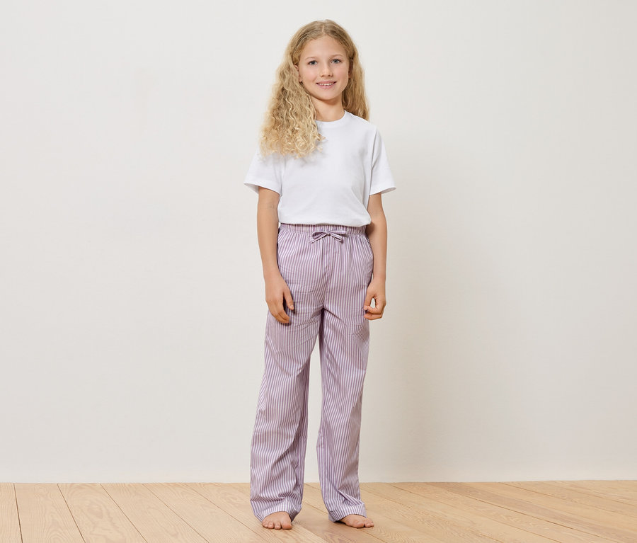 Dziewczynka z blond, kręconymi włosami w białym T-shircie i fioletowych spodniach w paski stoi boso na drewnianej podłodze na tle białej ściany.