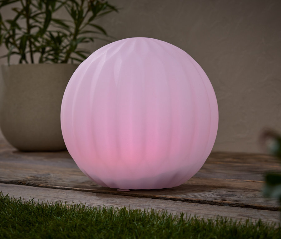 Na drewnianym stole stoi różowa lampa w kształcie kuli, obok doniczki z rośliną.