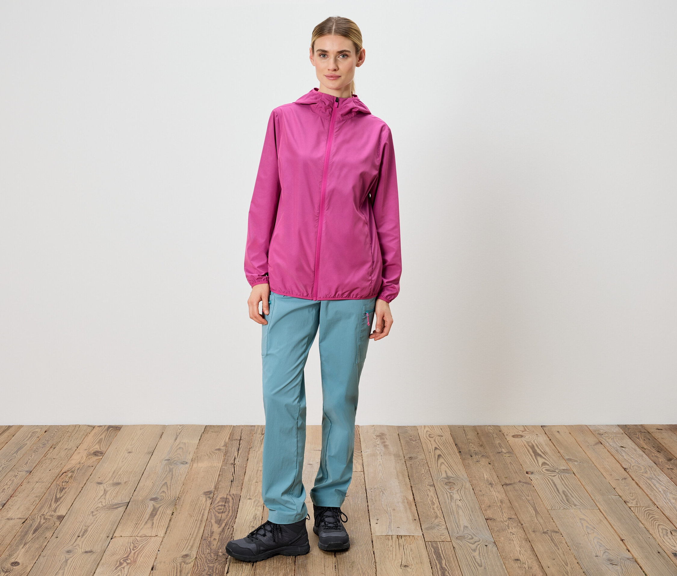 Kobieta stoi na tle białej ściany, ubrana w różową kurtkę, niebieskie spodnie i czarne buty.