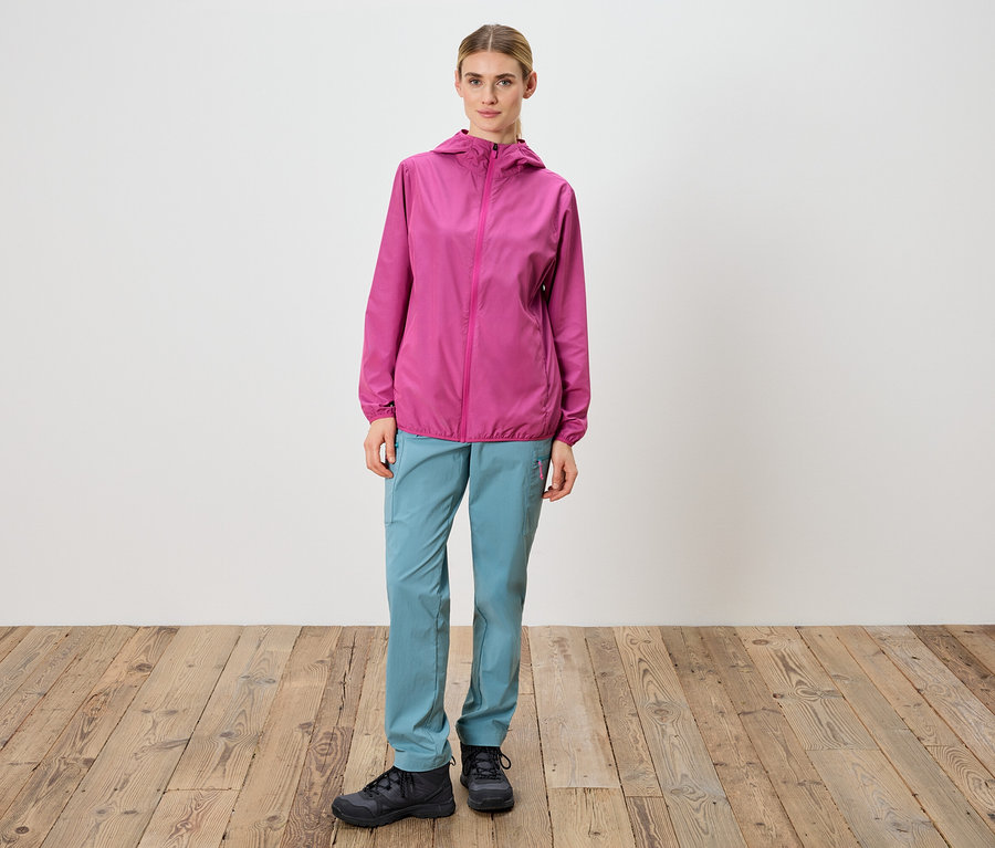 Kobieta stoi na tle białej ściany, ubrana w różową kurtkę, niebieskie spodnie i czarne buty.