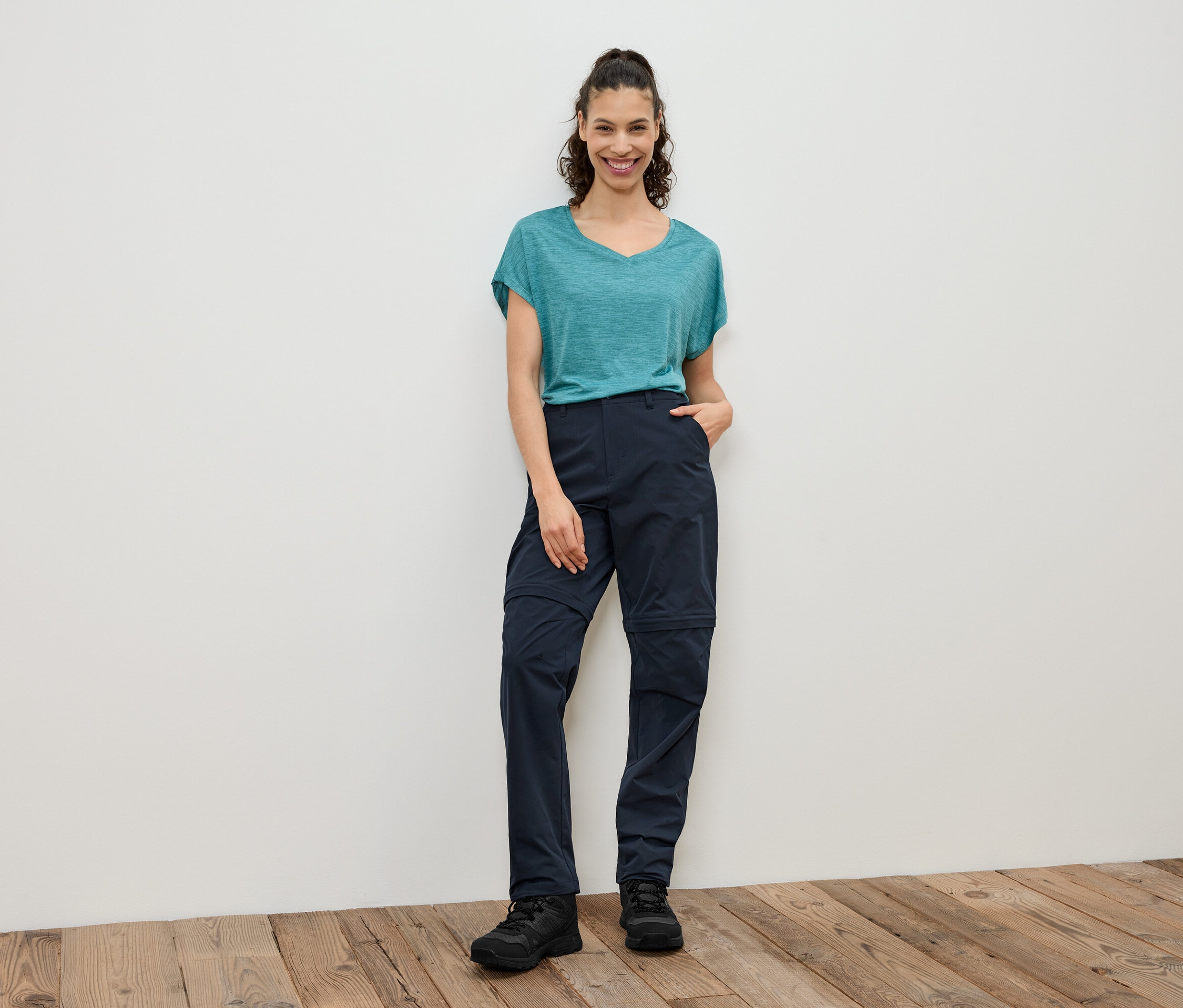 Kobieta stoi na tle białej ściany, ma na sobie turkusową koszulkę, granatowe spodnie z odpinanymi nogawkami i czarne buty.