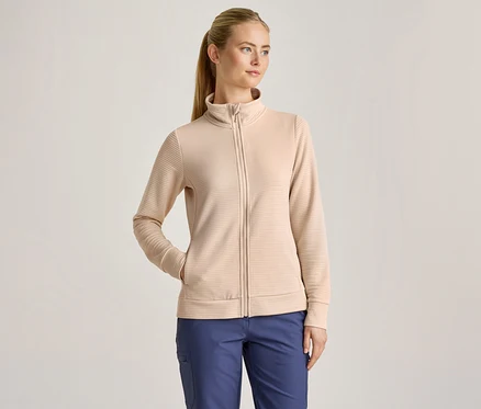 Kobieta z blond kucykiem ma na sobie beżową, prążkowaną kurtkę zapinaną na zamek i niebieskie spodnie, z jedną ręką w kieszeni kurtki.