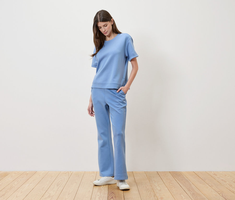 Kobieta o brązowych włosach ma na sobie jasnoniebieską koszulkę i pasujące spodnie, trzymając rękę w kieszeni.