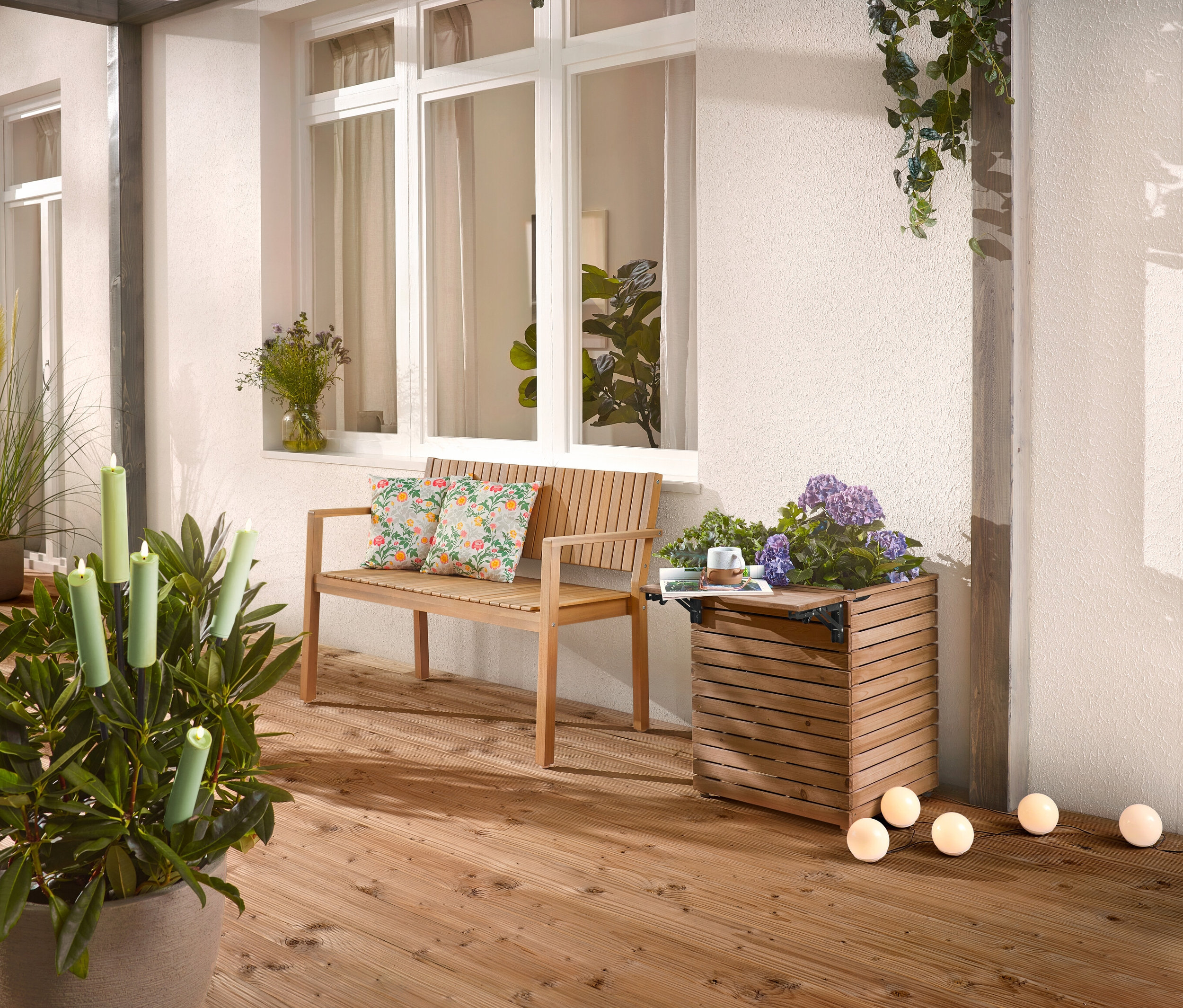 Na drewnianym tarasie stoi drewniana ławka z poduszkami, skrzynka na rośliny z kwiatami i roślina doniczkowa ze świecami.