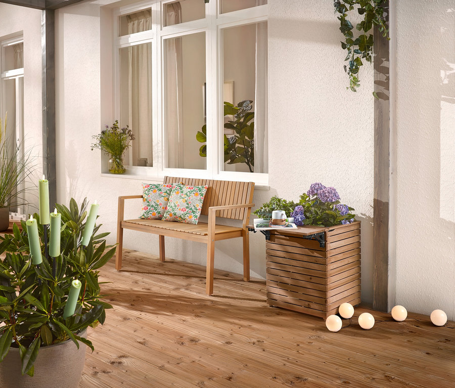 Na drewnianym tarasie stoi drewniana ławka z poduszkami, skrzynka na rośliny z kwiatami i roślina doniczkowa ze świecami.