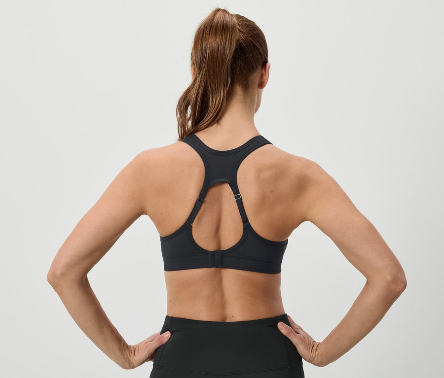 Widok z tyłu kobiety z brązowymi włosami upiętymi w koński ogon, ubranej w czarny biustonosz sportowy i legginsy, trzymającej ręce na biodrach.