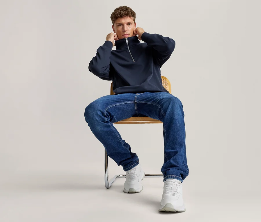 Mężczyzna siedzi na krześle, poprawiając kołnierz granatowej bluzy z krótkim zamkiem, ubrany w niebieskie dżinsy i białe sneakersy.