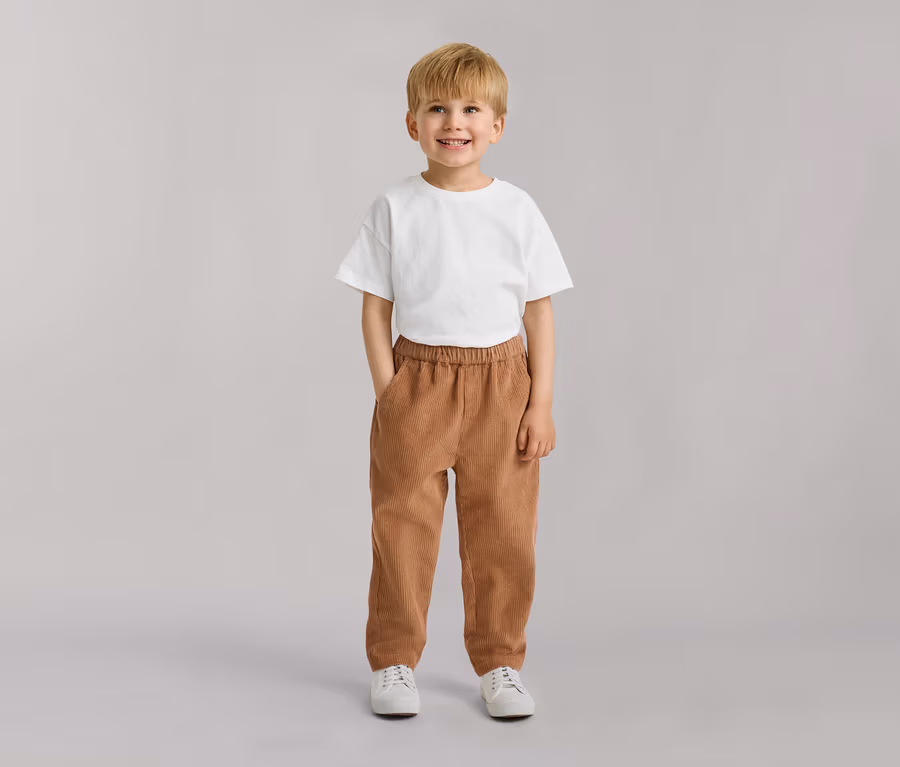 Uśmiechnięty mały chłopiec z blond włosami, w białym T-shircie, brązowych sztruksowych spodniach i białych trampkach, z rękami w kieszeniach.