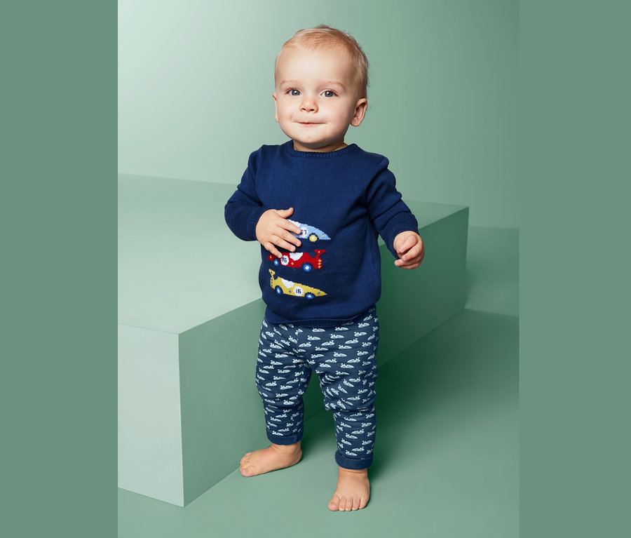 Dziecko stoi i ma na sobie niebieski sweter z samochodami i spodnie dresowe.