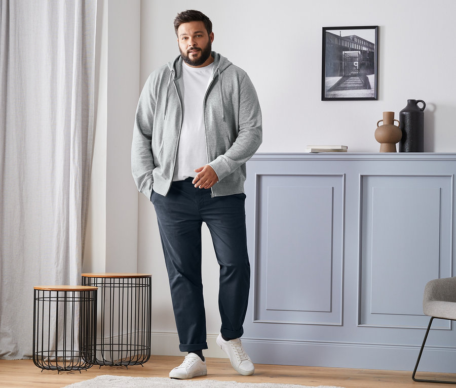 Mężczyzna stoi w szarej bluzie z dzianiny dresowej, białym t-shircie, niebieskich spodniach chino i białych tenisówkach. Widoczne są również skarpetki z wełny merino, 4 pary, czarne, granatowe, antracytowe i szare.