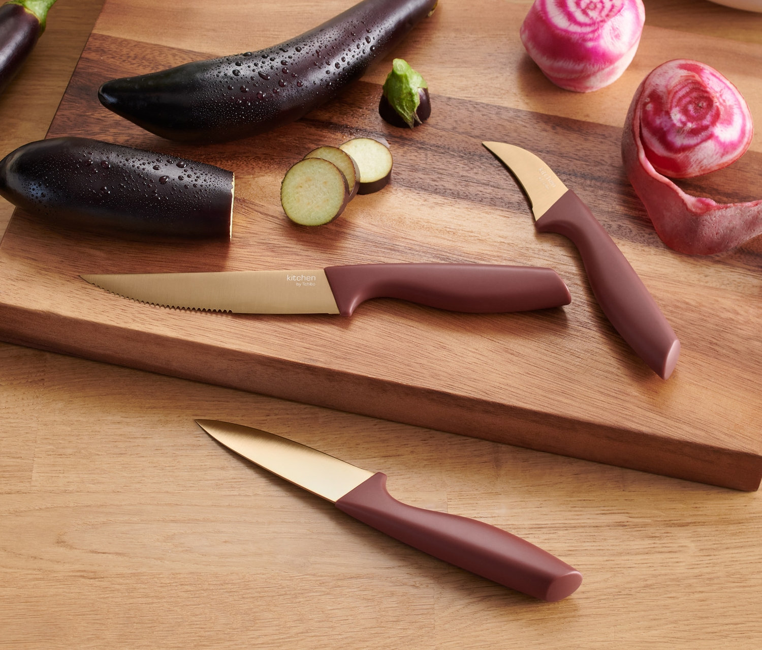 Na drewnianej desce leżą trzy bakłażany, pokrojone buraki i trzy noże kuchenne.