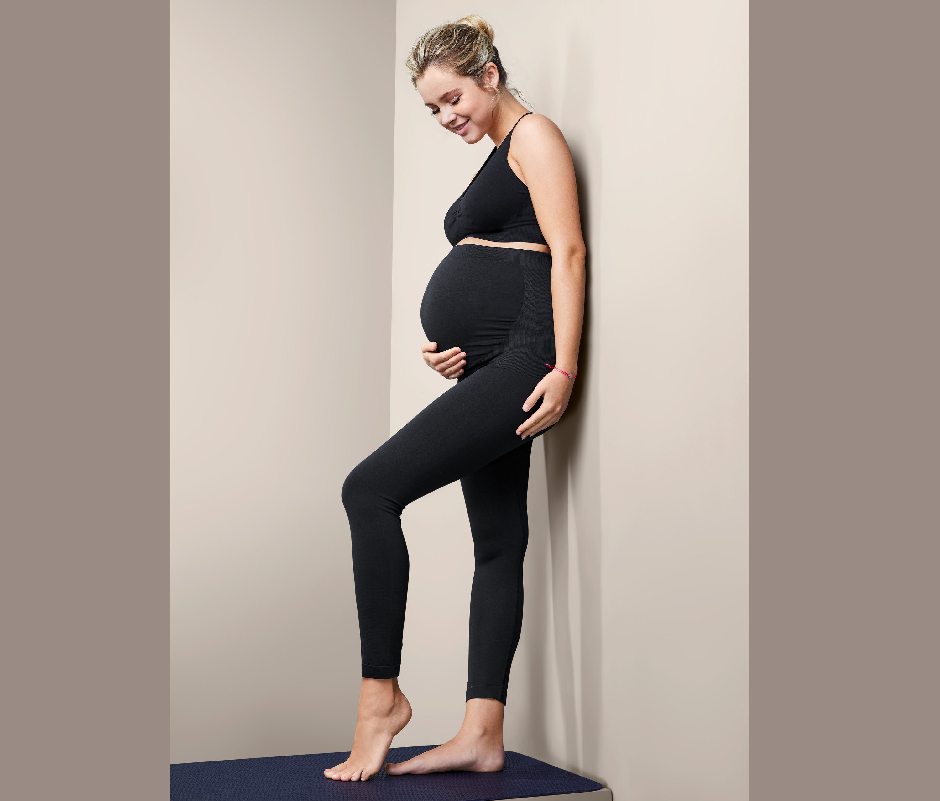 Kobieta w ciąży w czarnych bezszwowych legginsach ciążowych stoi bokiem przy ścianie.