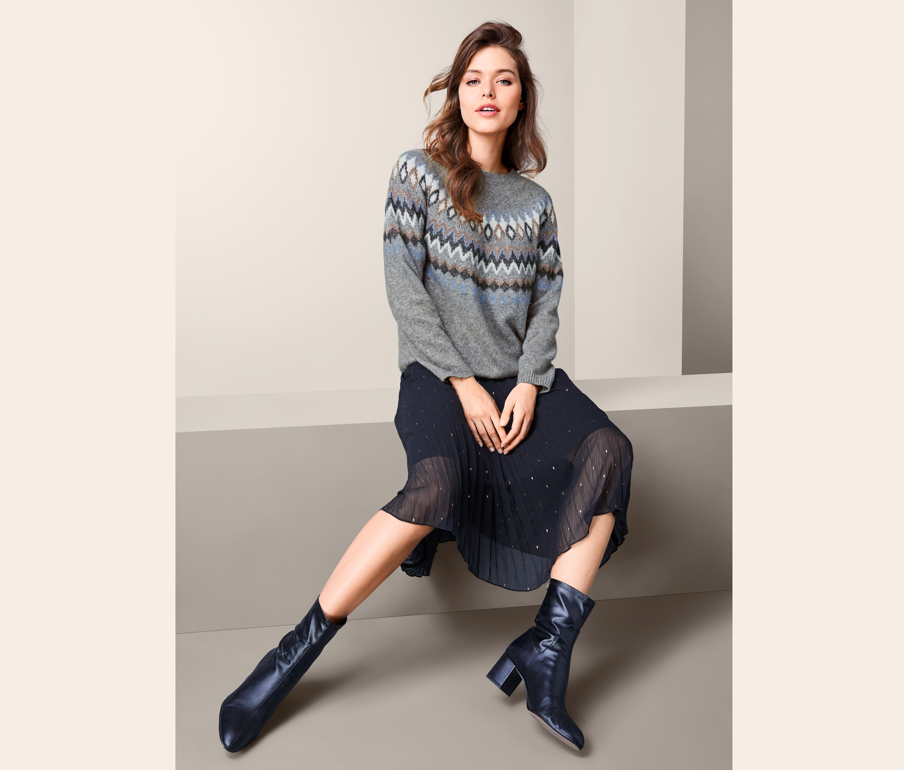 Kobieta siedzi i ma na sobie sweter damski z dzianiny z norweskim wzorem, plisowaną spódnicę i skórzane buty.