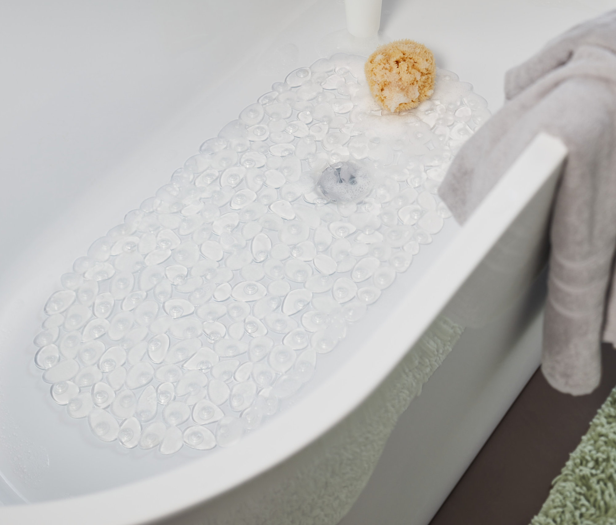 W wannie leży przezroczysta mata do wanny. Na krawędzi wanny wisi szary ręcznik, a obok wanny leży zielony dywan.