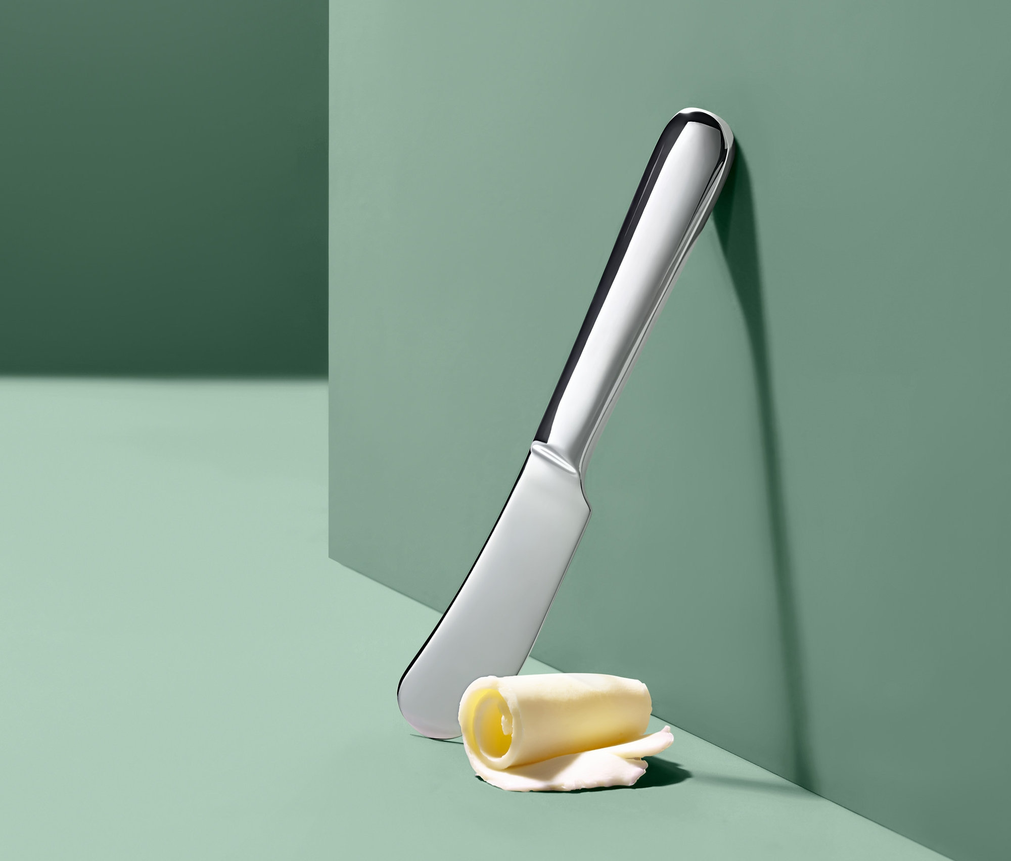 Srebrny nóż do masła i zwinięta rolka masła oparte o zieloną ścianę.