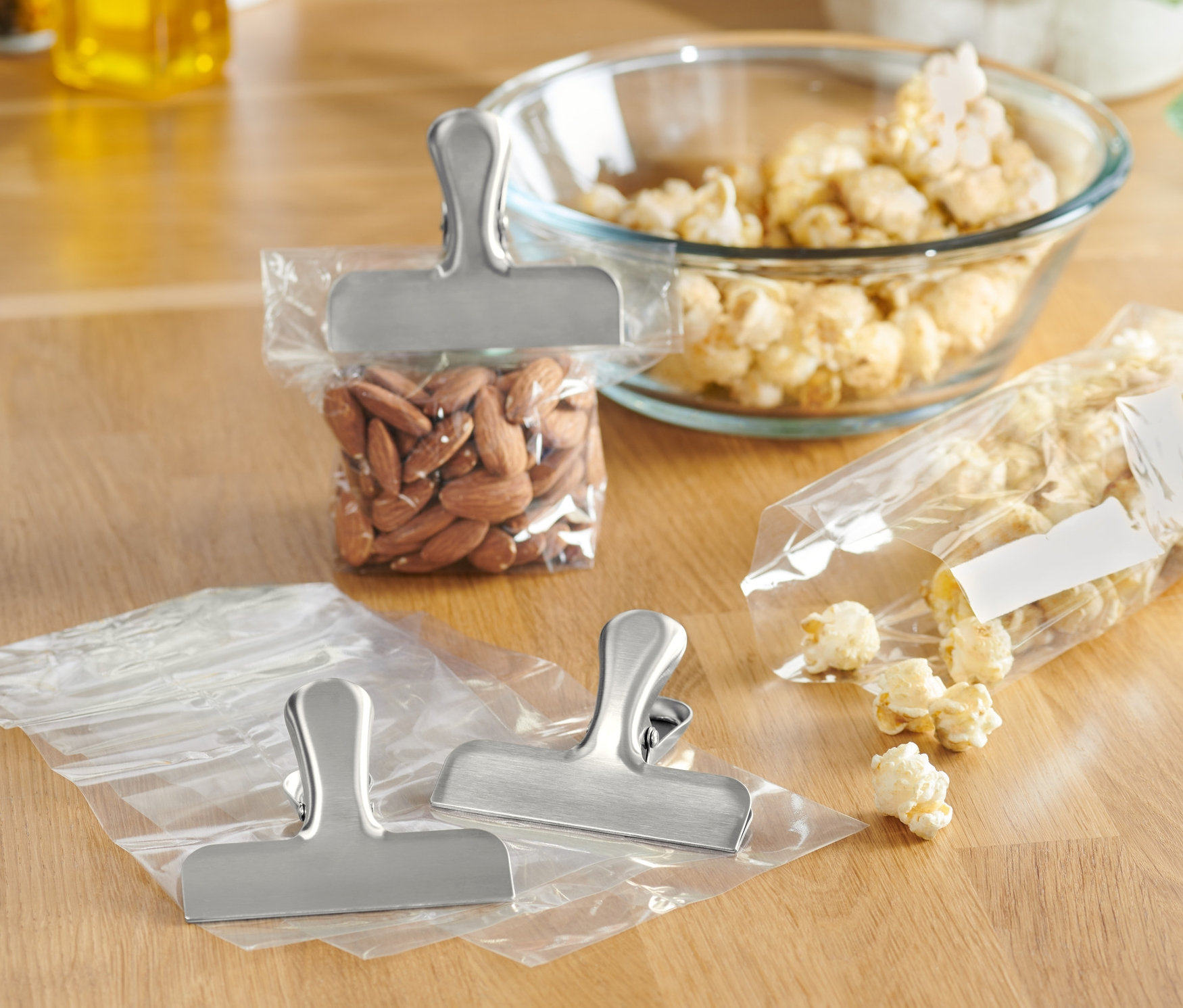 Na drewnianym stole leżą 3 klipsy ze stali nierdzewnej, zamykające torebki z migdałami i popcornem.