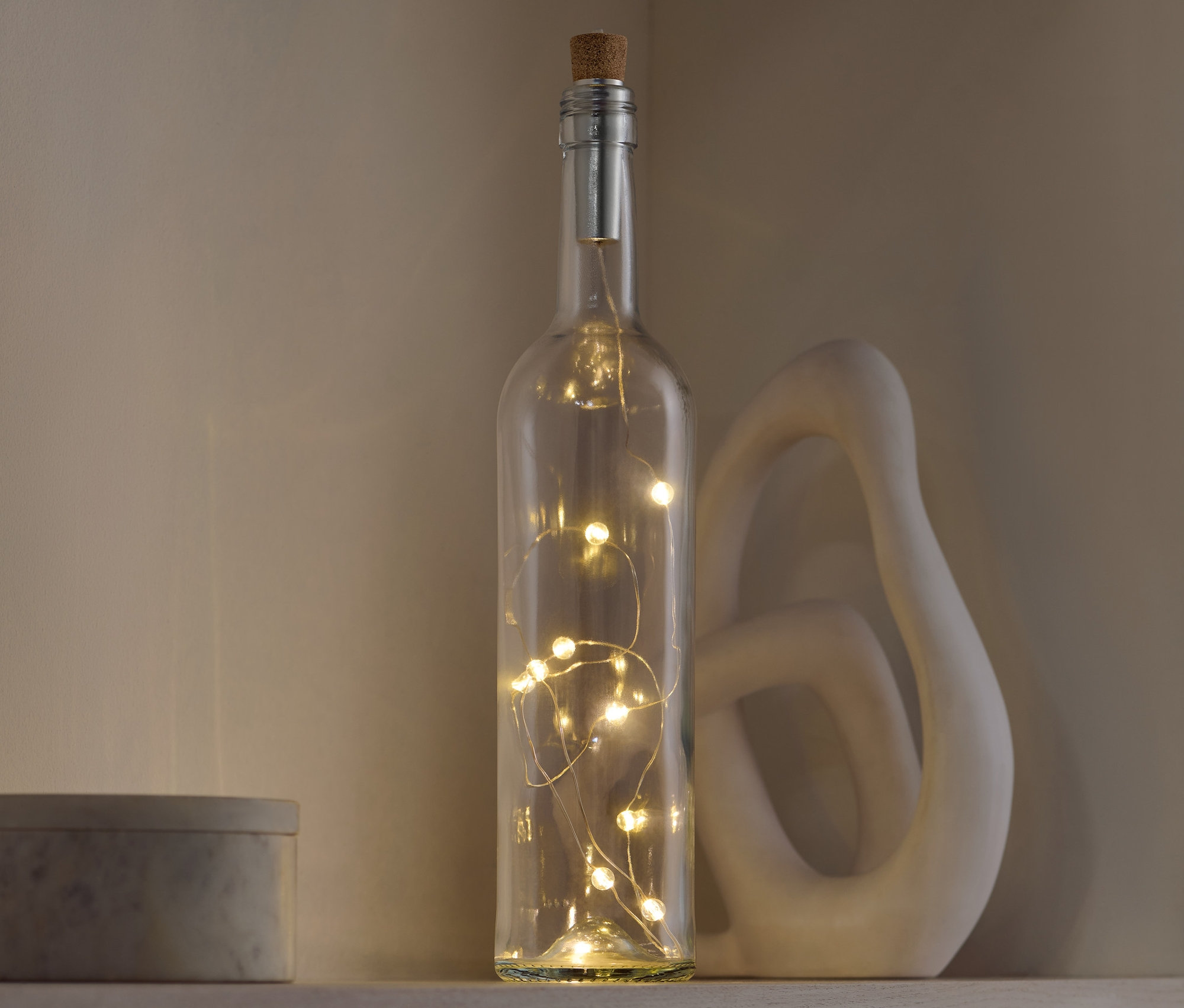 Dekoracyjna szklana butelka z korkiem, wewnątrz której znajdują się światełka, stoi na półce obok ozdobnej figurki i szarego pudełka.
