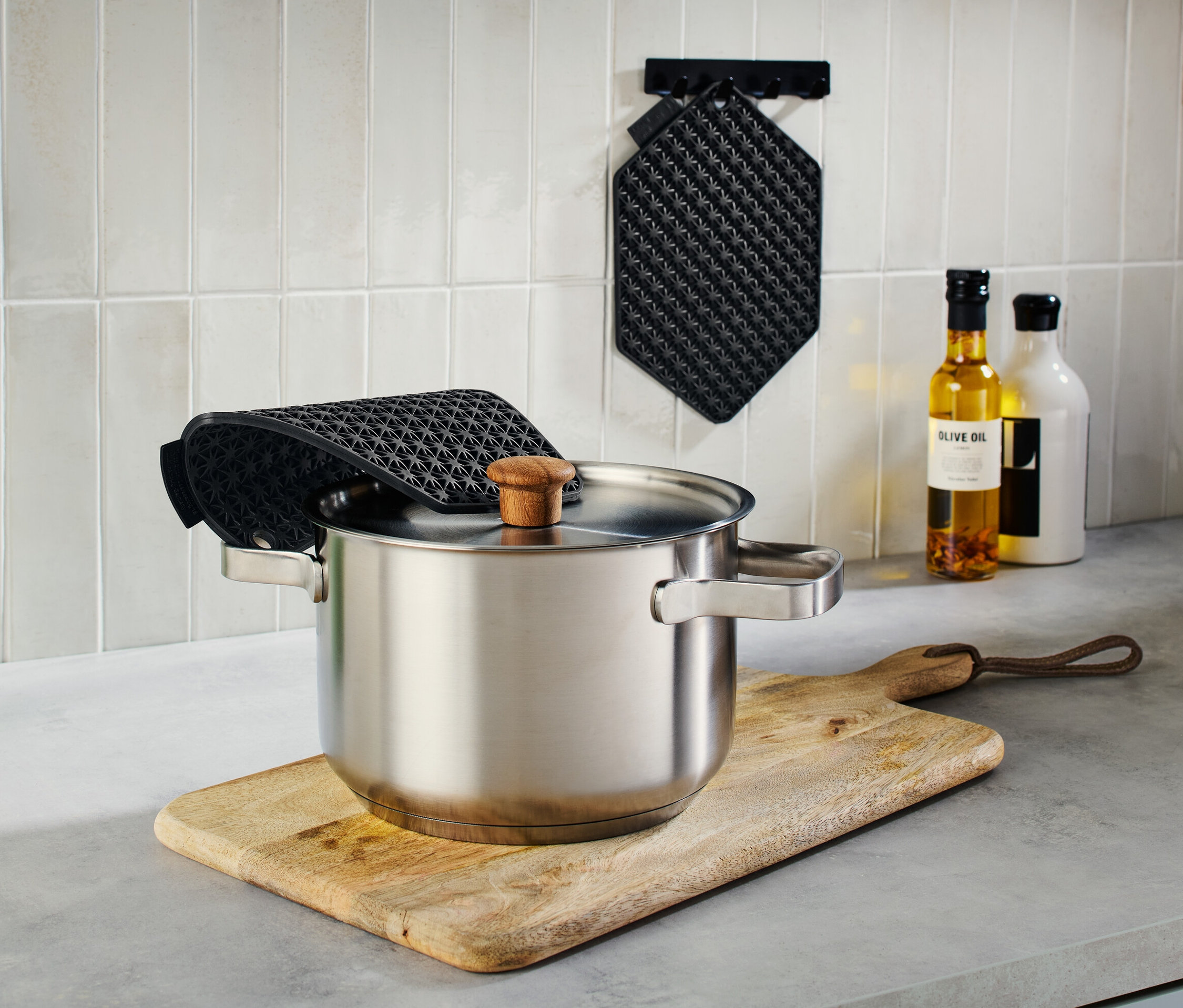 Garnek ze stali nierdzewnej z pokrywką na drewnianej desce do krojenia, obok zielony dzbanek z przyborami kuchennymi i czarne podkładki kuchenne 3 w 1.
