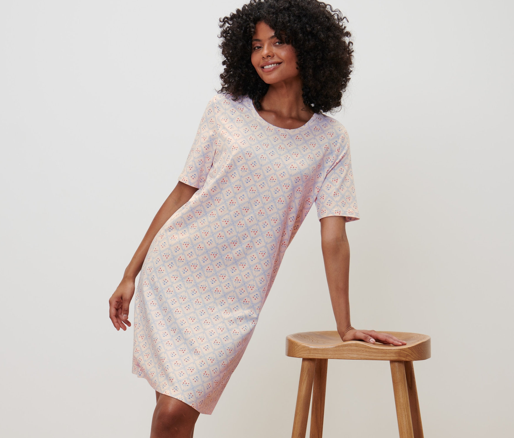 Kobieta pozuje w koszuli nocnej z krótkim rękawem, opierając się o drewniany stołek.
