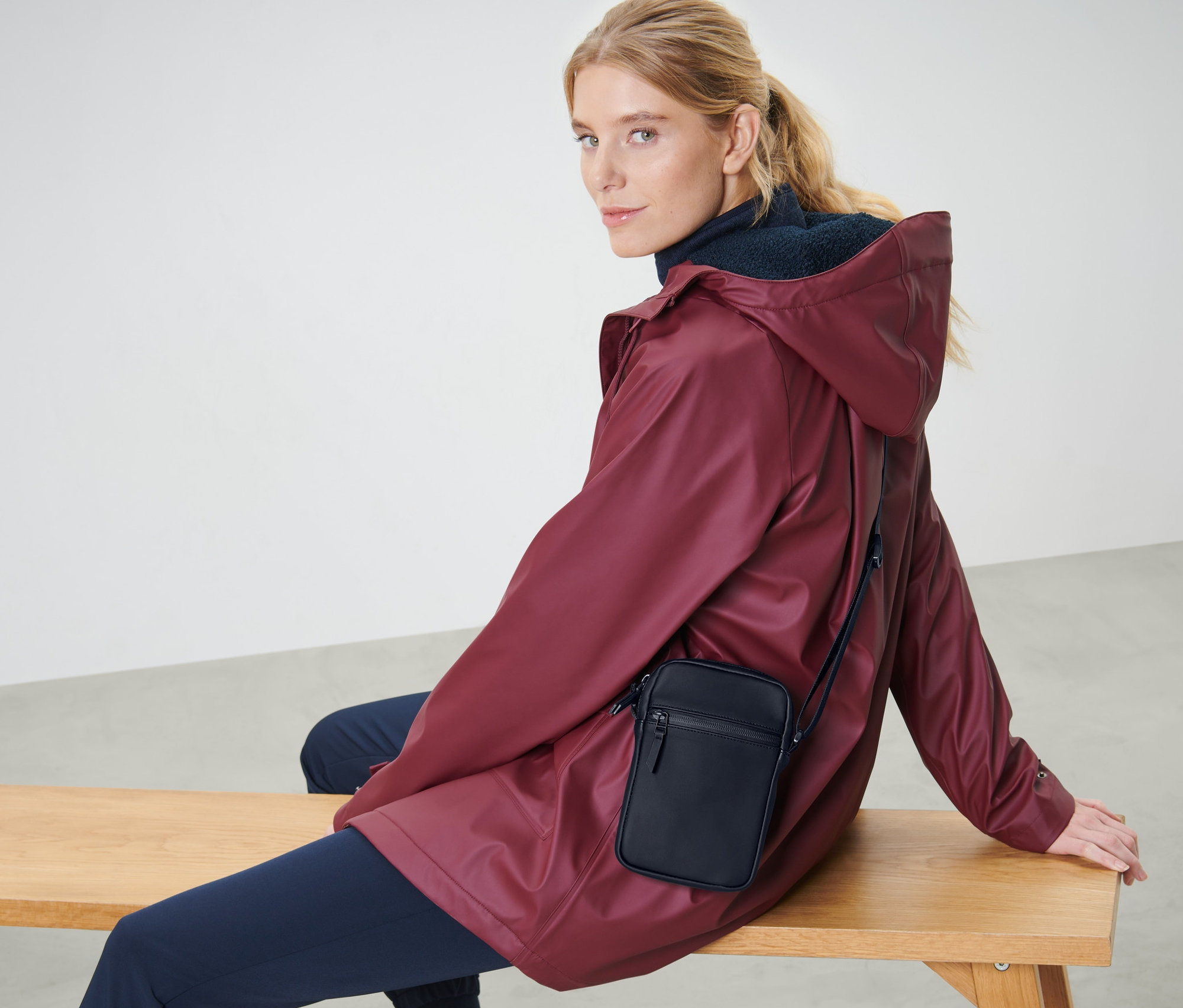 Blondwłosa kobieta siedzi na ławce w bordowej kurtce i z czarną przeciwdeszczową torbą na ramię.