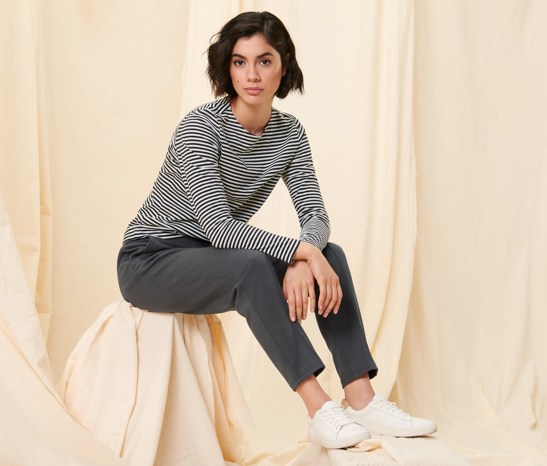 Kobieta siedzi na stołku pokrytym materiałem i ma na sobie czarną bluzkę z długimi rękawami w paski oraz spodnie dresowe z kantem.