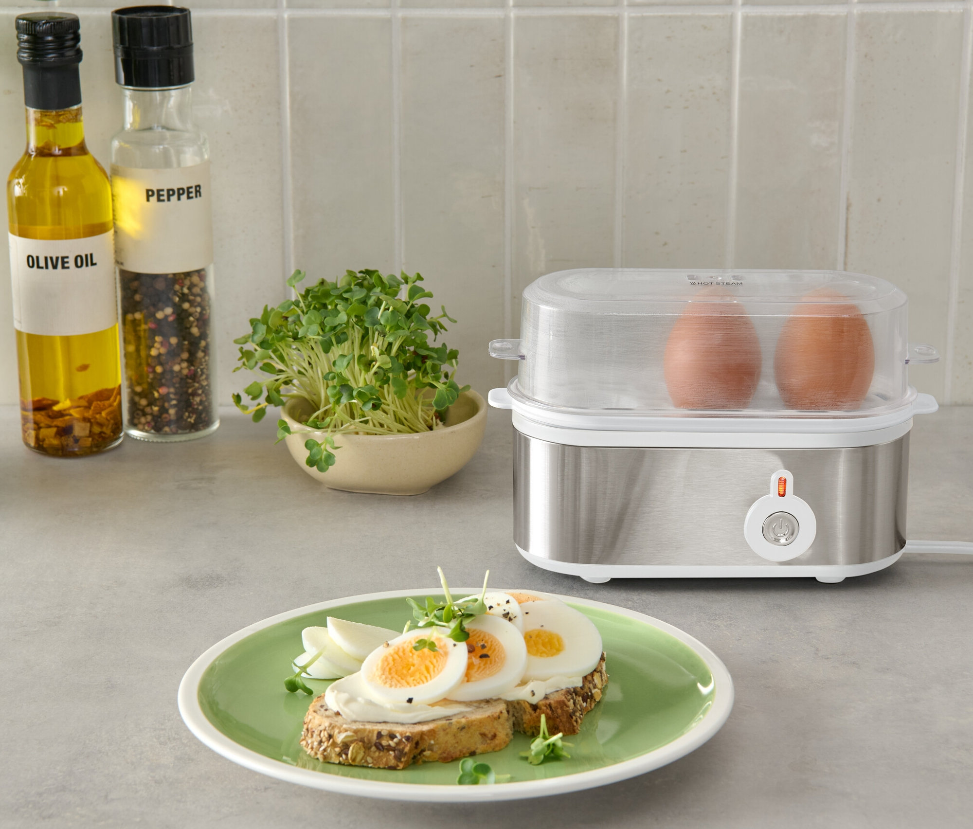 Dwa brązowe jajka w srebrnym jajowarze z przezroczystą pokrywką, obok talerza z chlebem i jajkami.