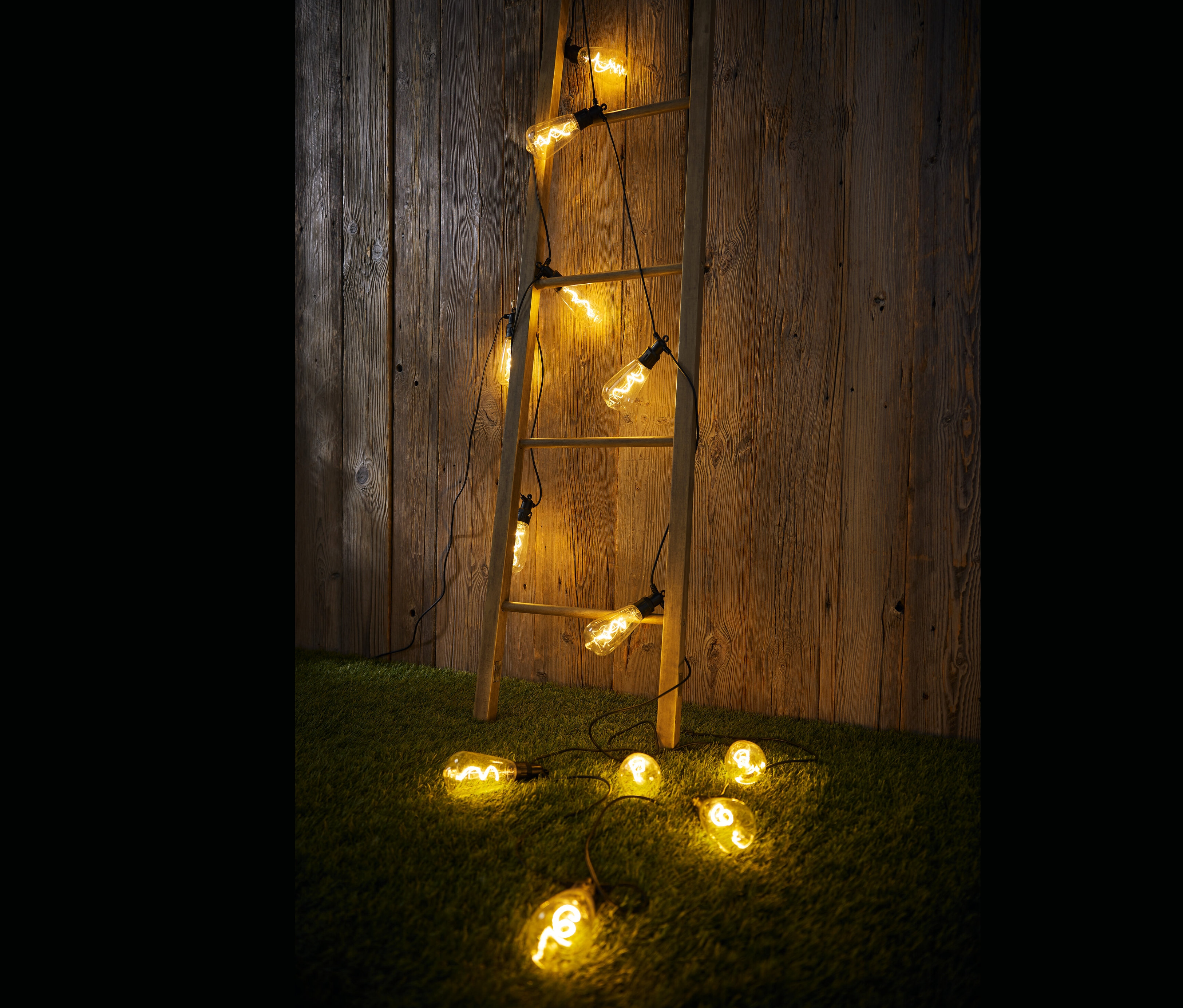 Drewniana drabina oparta o drewnianą ścianę, na której i na ziemi leży łańcuch świetlny LED do użytku na zewnątrz.