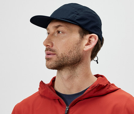 Mężczyzna w pomarańczowej kurtce ma na sobie czapkę z daszkiem i patrzy w lewo.