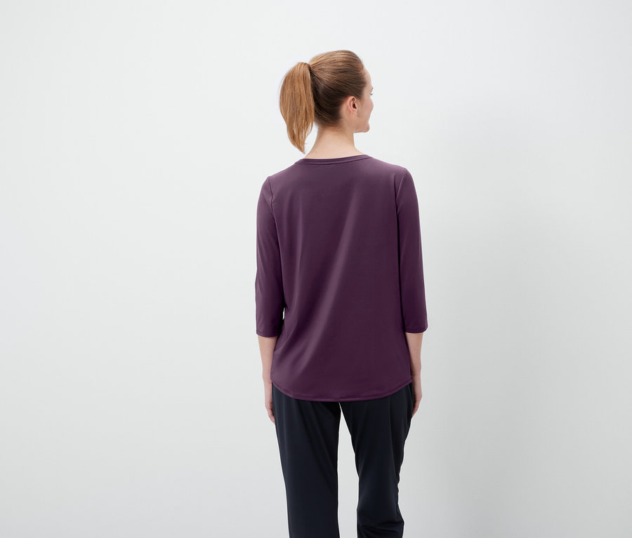 Widok z tyłu na kobietę z brązowymi włosami związanymi w kucyk, ubraną w fioletową koszulkę z rękawami trzy czwarte i czarne spodnie.