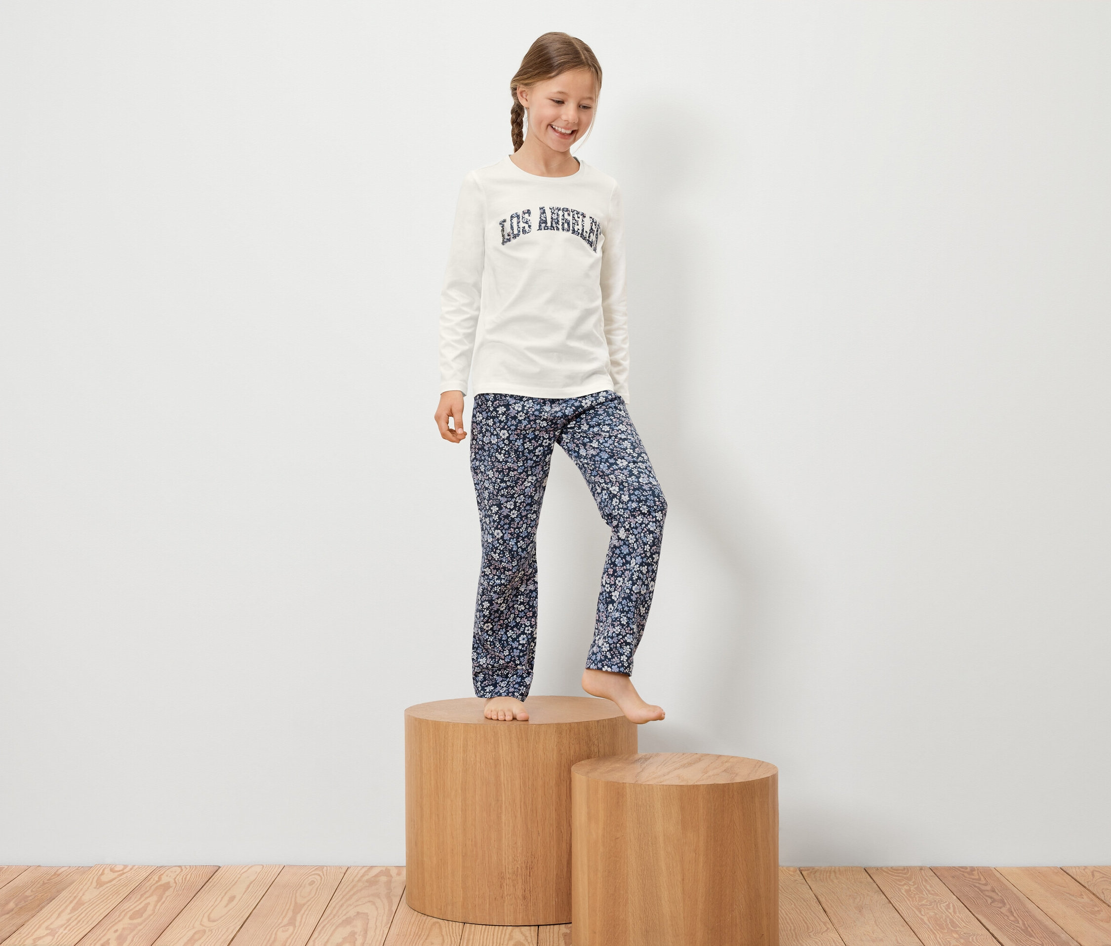 Dziewczynka z warkoczem stoi na drewnianym cokole w białej koszulce z napisem i kwiecistych spodniach.