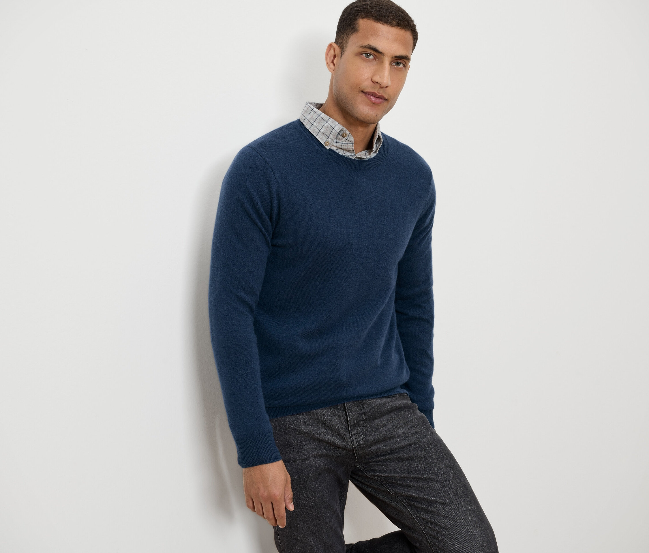 Mężczyzna oparty o ścianę w niebieskim swetrze i szarych wzorzystych spodniach.