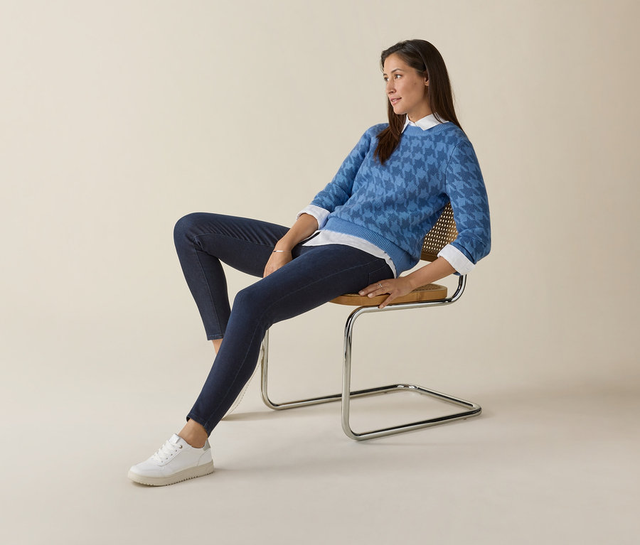 Kobieta siedzi na krześle, ubrana w niebieski sweter i białe sneakersy Tom Tailor.