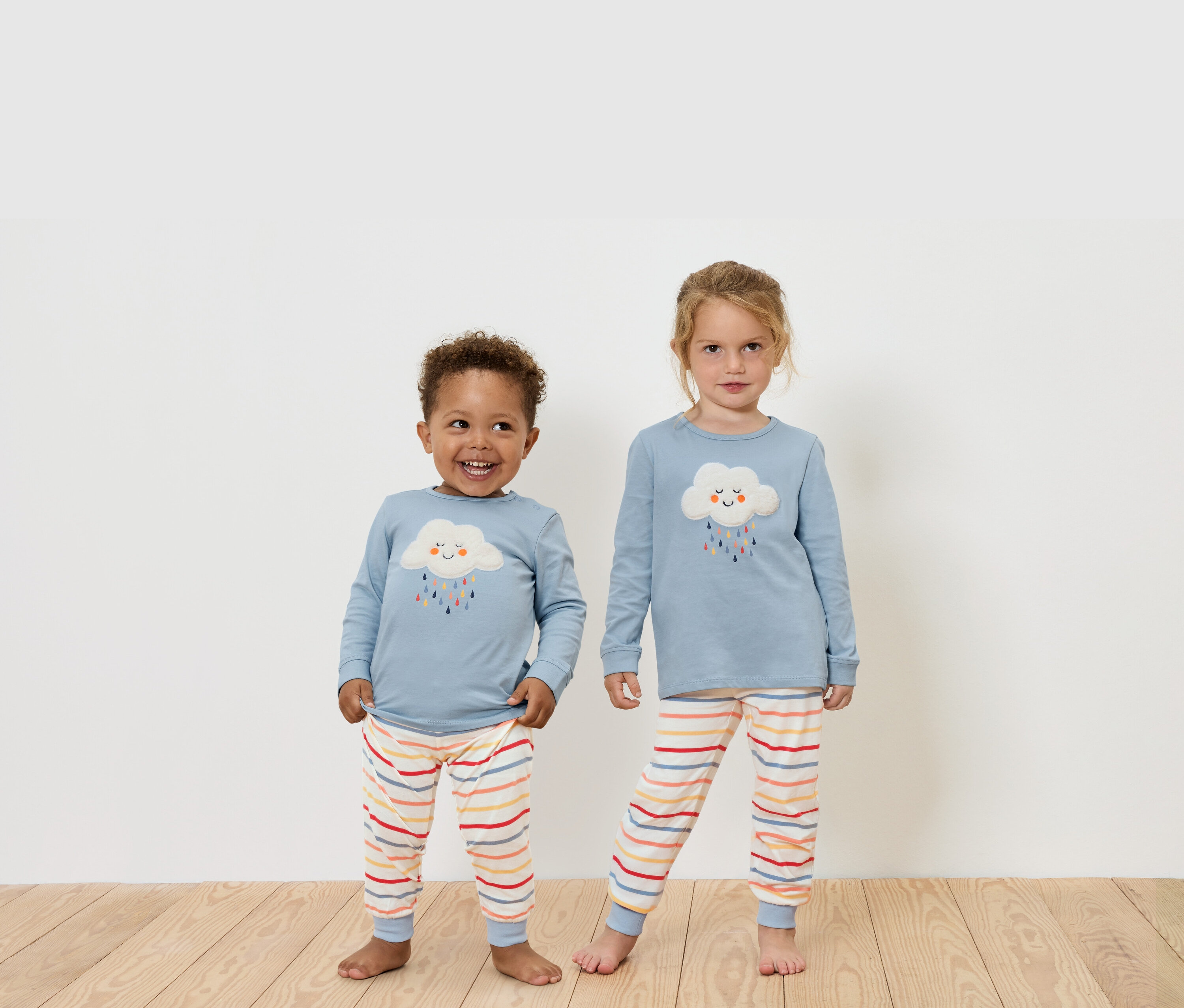 Dwoje dzieci stoi na drewnianej podłodze, ubrani w niebieskie koszulki z długim rękawem i spodnie w paski.
