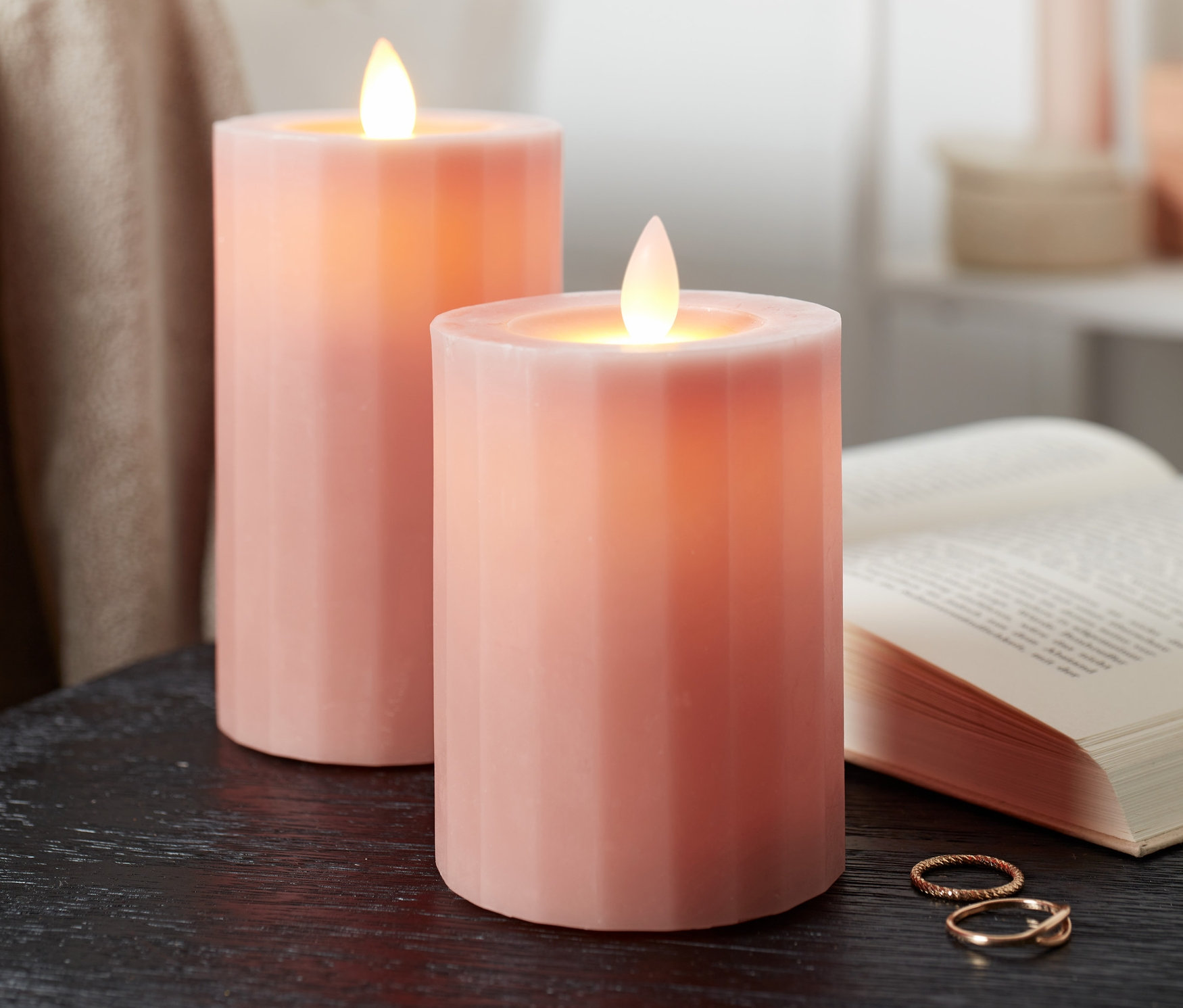 Dwie świece LED z prawdziwego wosku stoją na czarnym stole obok otwartej książki.