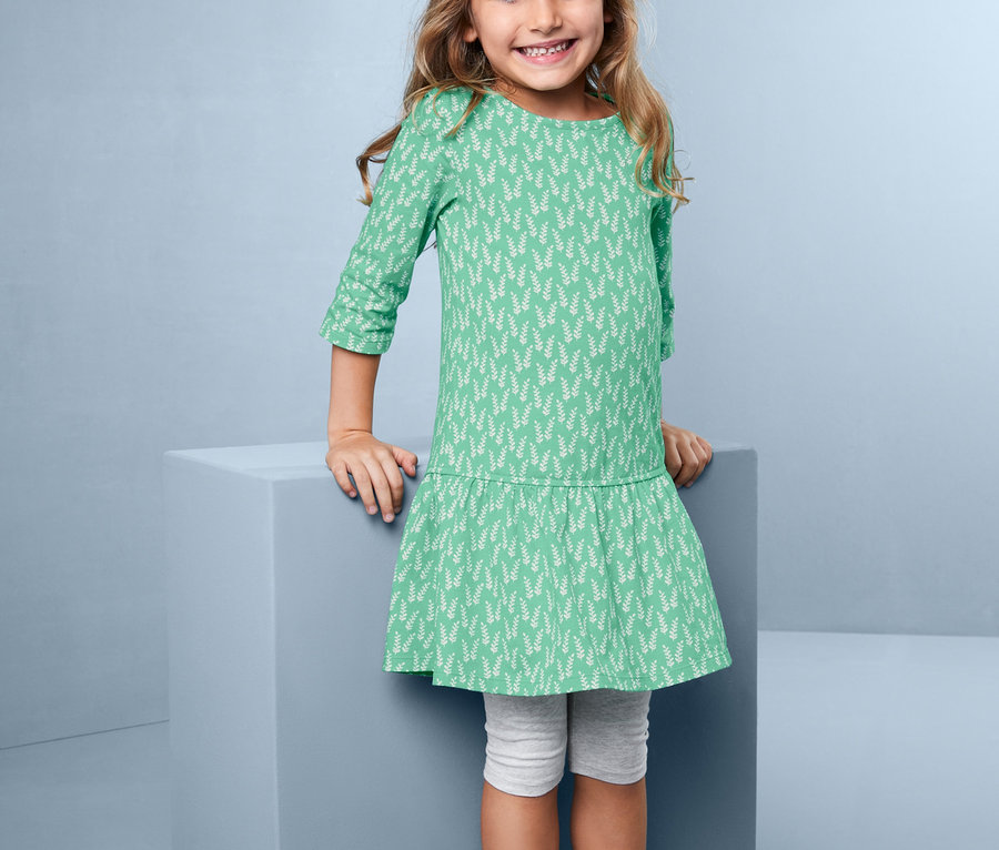 Uśmiechnięta dziewczynka pozuje w sukience dżersejowej z kwiatowym nadrukiem na całej powierzchni.