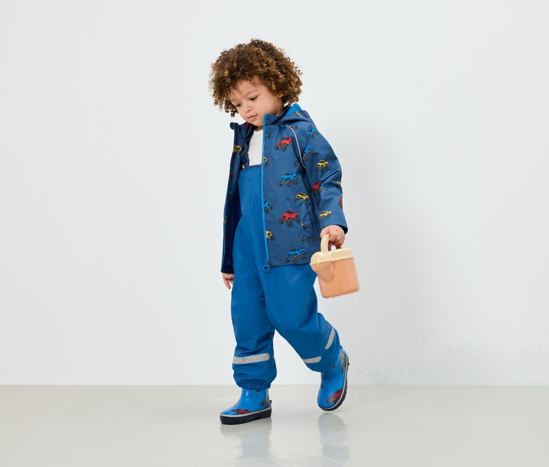 Chłopiec w niebieskich spodniach na szelkach, kurtce z nadrukiem samochodów i niebieskich kaloszach trzyma beżową zabawkę.