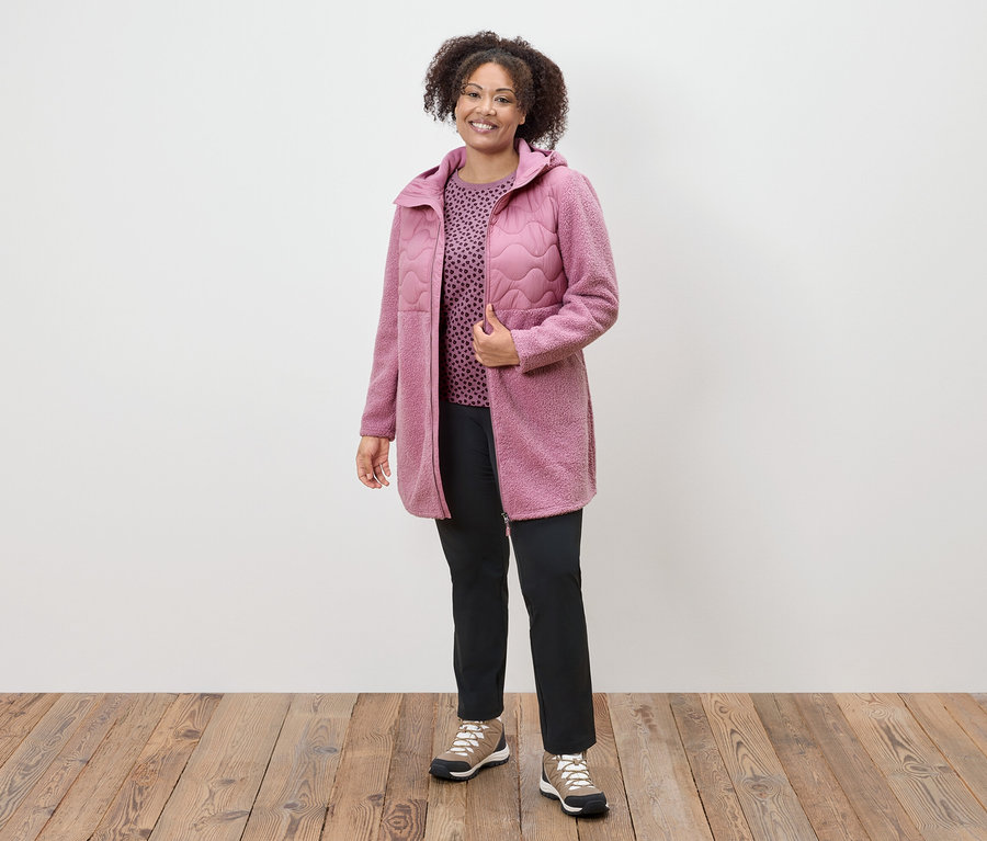 Kobieta w różowym płaszczu, wzorzystej bluzce i czarnych spodniach stoi na drewnianej podłodze.