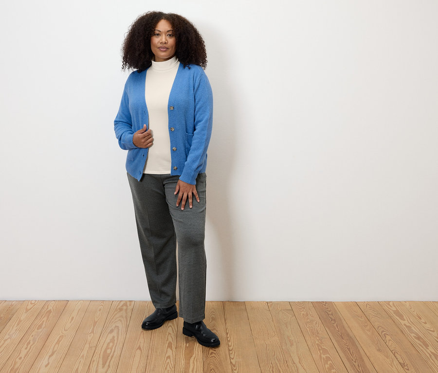 Kobieta stojąca na drewnianej podłodze ma na sobie wygodne spodnie dżersejowe w kratkę, biały golf i niebieski kardigan.