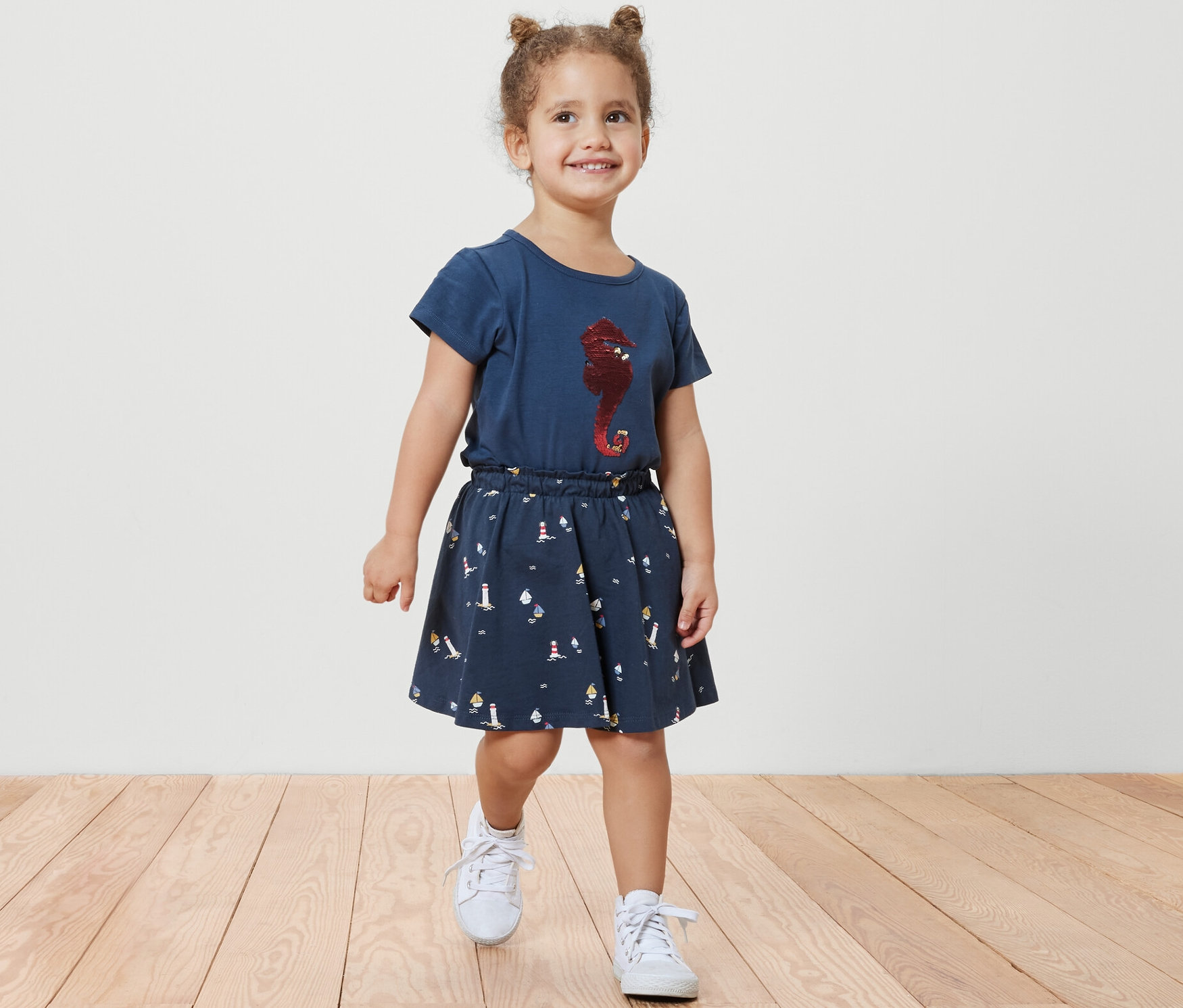 Uśmiechnięta dziewczynka idzie po drewnianej podłodze, ubrana w spódnicę z dżerseju ze spodenkami, w stylu marynistycznym, oraz niebieską koszulkę z nadrukiem konika morskiego.