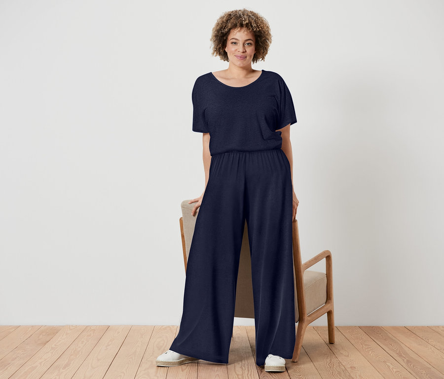 Kobieta o kręconych włosach ma na sobie granatowy kombinezon i białe buty, stoi obok krzesła z drewnianą ramą.