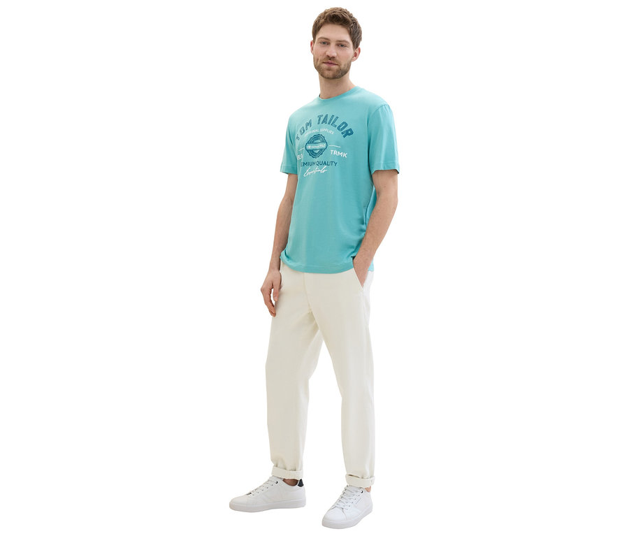 Mężczyzna pozuje w koszulce z logo Tom Tailor, meadow teal, beżowych spodniach i białych sneakersach.
