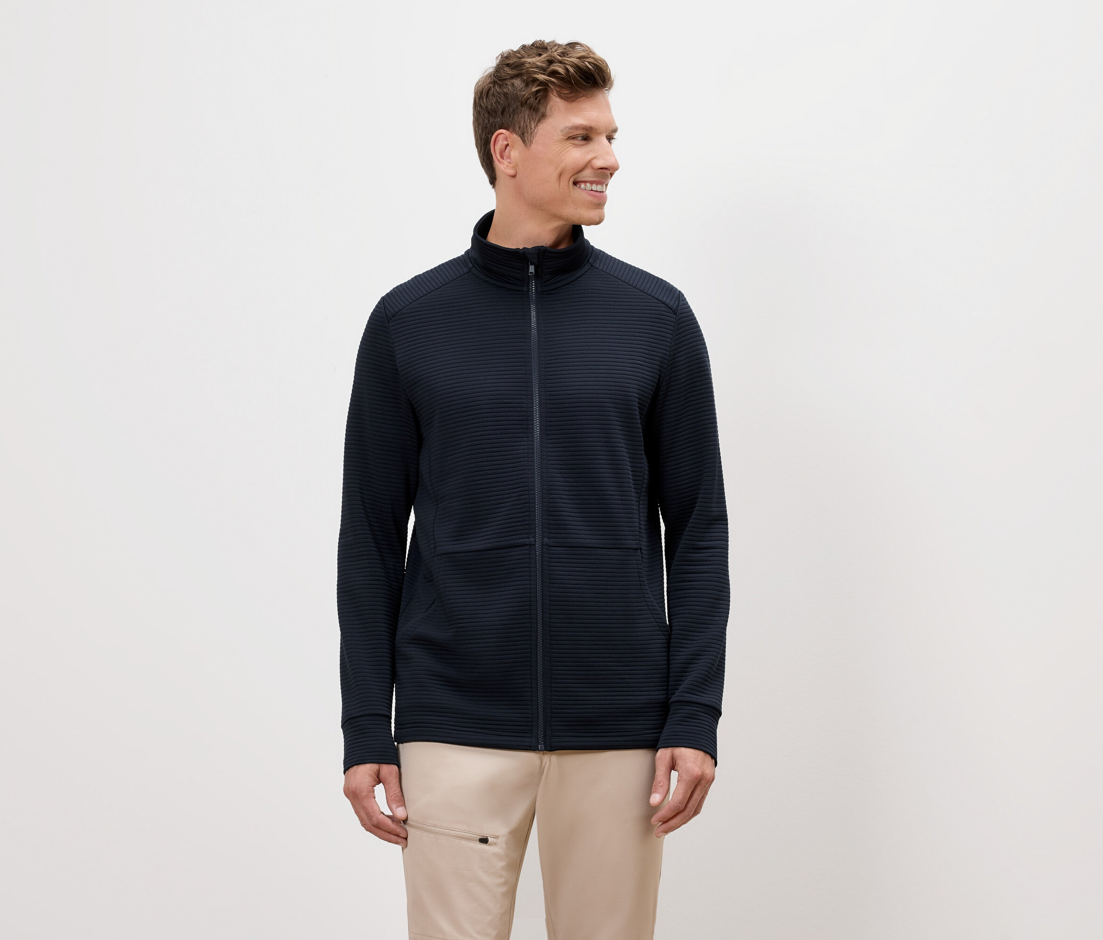 Mężczyzna o blond włosach ma na sobie granatową bluzę z zamkiem błyskawicznym i beżowe spodnie.