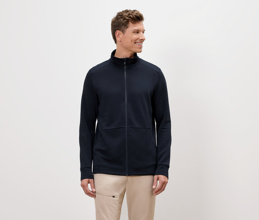 Mężczyzna o blond włosach ma na sobie granatową bluzę z zamkiem błyskawicznym i beżowe spodnie.