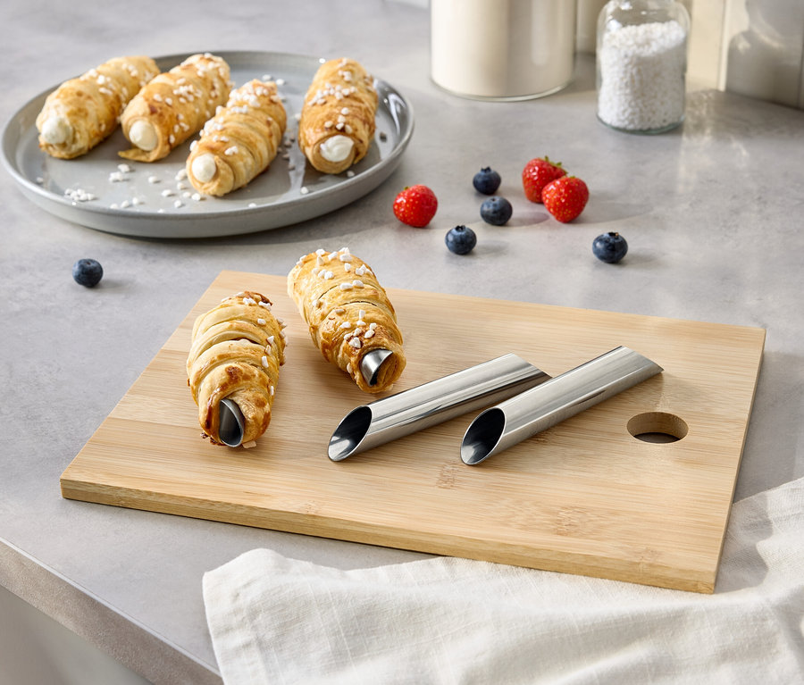 Na stole leżą rurki z kremem na talerzu i desce do krojenia, wraz z jagodami, cukrem i metalowymi foremkami.